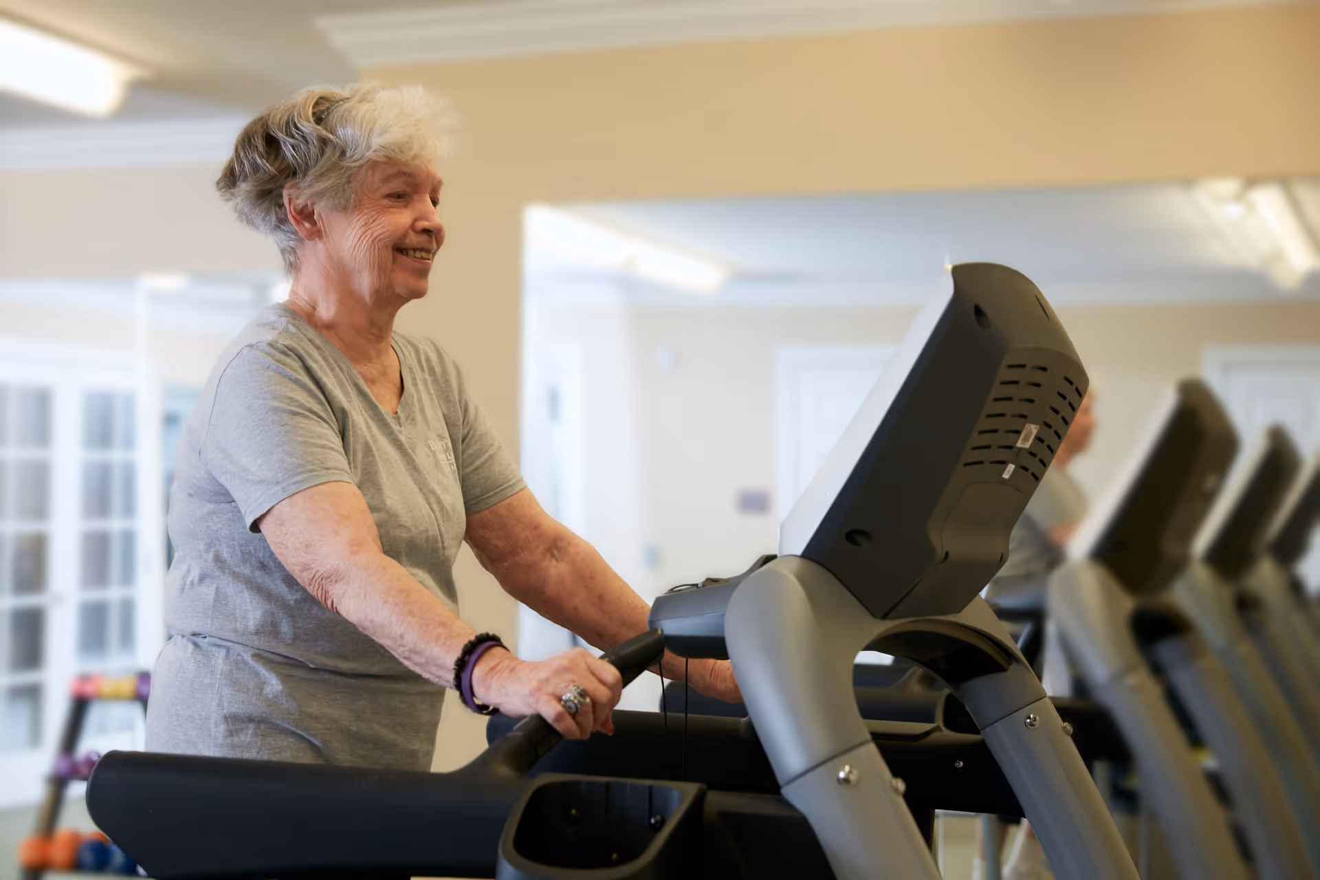An elderly woman with short gray hair is smiling while walking on a treadmill in a fitness room with light-colored walls and large windows.
