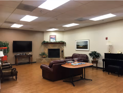 Communal lounge with leather sofas, a fireplace, a wall-mounted TV, a piano, and a central table in a senior living facility.
