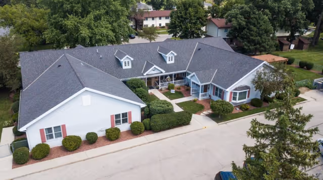 Aerial view of a single-story assisted living facility building with a dark gray roof, white exterior walls, and red window shutters. The building is surrounded by neatly trimmed bushes and trees, with a paved road in front and additional residential buildings in the background.