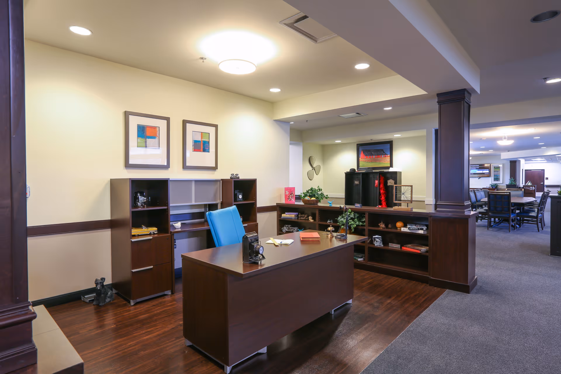 A well-lit office area in Orchard Grove Senior Living featuring a dark wooden desk with a blue office chair, shelves with decorative items, framed abstract artwork on the wall, and an open view into a common area with tables and chairs.