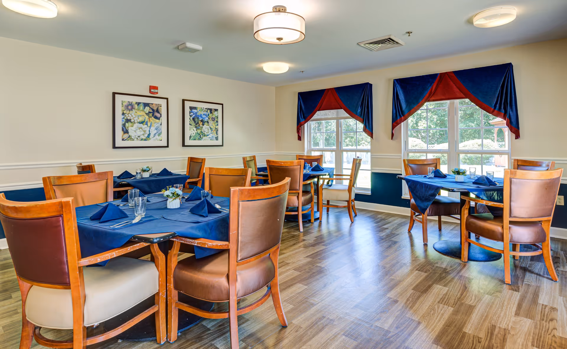 A bright dining room with wooden floors and several tables covered with blue tablecloths and matching blue napkins. Each table is surrounded by wooden chairs with cushioned seats. Two large windows with blue and red valances allow natural light to fill the room. The walls are light-colored with two framed floral paintings hanging on one side.