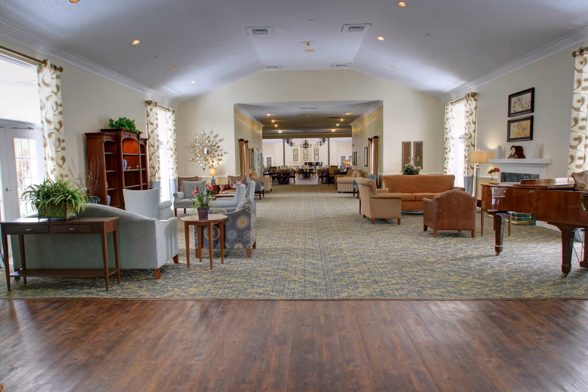 A spacious and well-lit senior living common area with multiple seating arrangements including armchairs and sofas. The room features large windows with patterned curtains, a wooden bookshelf, a grand piano, and decorative plants. The flooring transitions from wood to carpet, and the room extends into a dining area in the background.