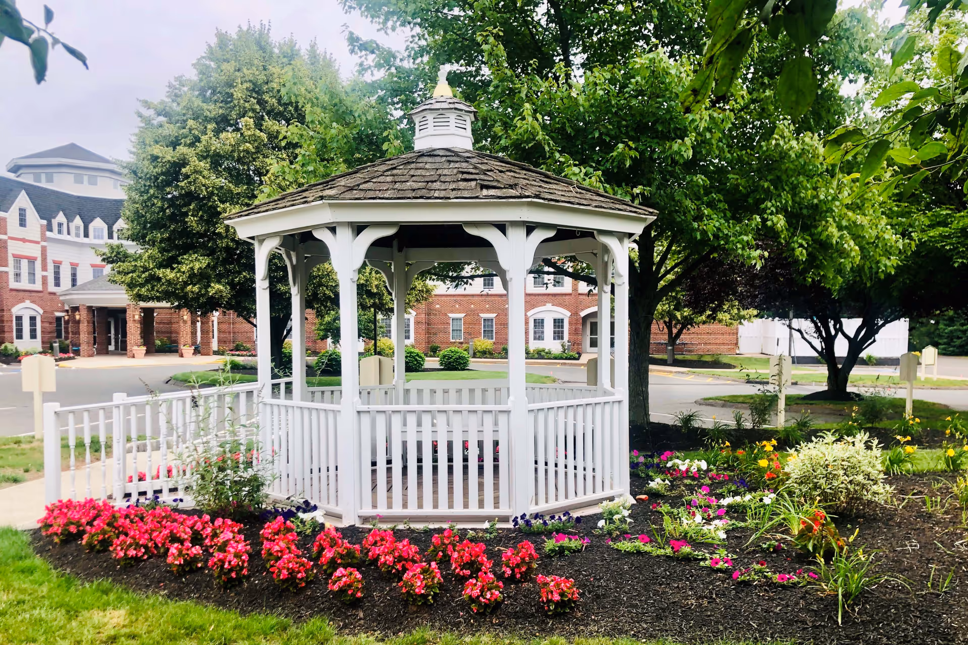A white wooden gazebo with a shingled roof is surrounded by colorful flower beds and green trees. In the background, there is a large brick building with white-trimmed windows and a covered entrance.