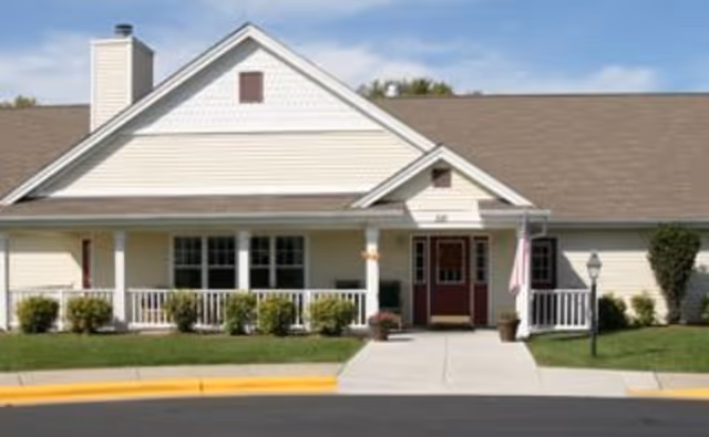 Front exterior view of a single-story building with beige siding, a brown shingled roof, a covered porch with white railings, shrubs along the front, and a concrete walkway leading to a red front door with an American flag on the right side.