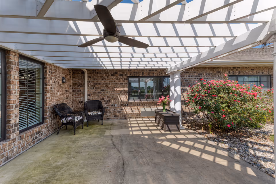Outdoor patio area with a white pergola casting shadows on the concrete floor. Two black wicker chairs with cushions are placed against a brick wall with windows. A ceiling fan is mounted under the pergola. There is a small table with a vase of flowers and a large bush with pink flowers nearby.
