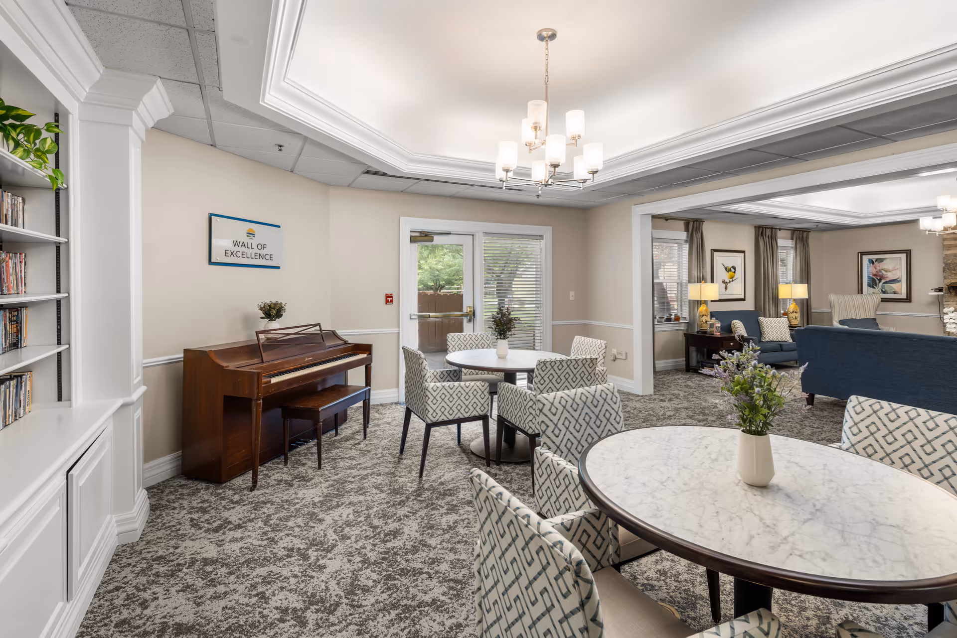 Bright senior living common room with round marble-top tables and patterned chairs, a piano and bookshelf, and a seating area under chandeliers.