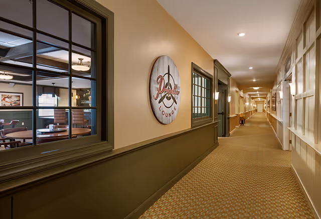 Carpeted interior hallway in a senior living facility with windows into a lounge and a round wall sign reading 'Lounge'.