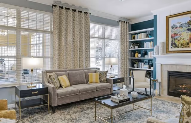 Bright living room with a gray sofa, coffee table, fireplace, built-in bookshelves and large windows with curtains.