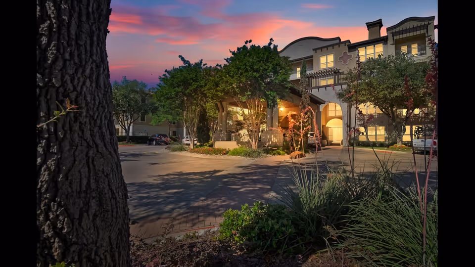 Front entrance of a residential building at dusk with lit windows, landscaped grounds, and a large tree trunk in the foreground.