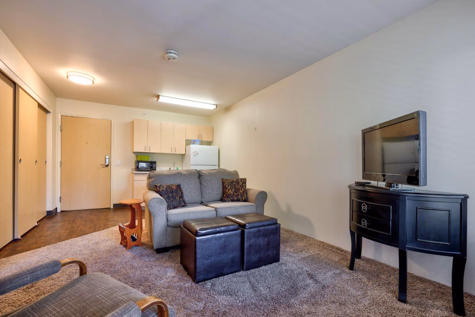 A small living area with a gray loveseat with two patterned pillows, two black ottomans in front of it, a wooden side table, and a TV on a black console table. The room has beige walls, carpeted floor, and a kitchenette with light wood cabinets, a white refrigerator, and a microwave in the background.