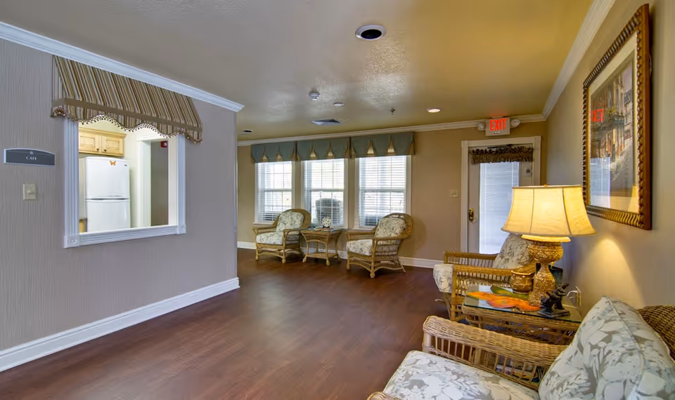 A cozy sitting area in a senior living facility with wicker chairs and a wicker sofa with floral cushions, a side table with a lamp, and a framed picture on the wall. There are three large windows with valances letting in natural light and a door with an exit sign above it. A small serving window labeled 'Cafe' looks into a kitchen area with a refrigerator.