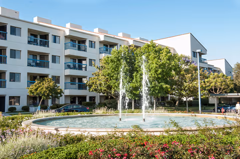 Exterior view of a multi-story senior living facility with balconies, surrounded by trees and greenery. In the foreground, there is a circular fountain with water jets and landscaped flower beds.