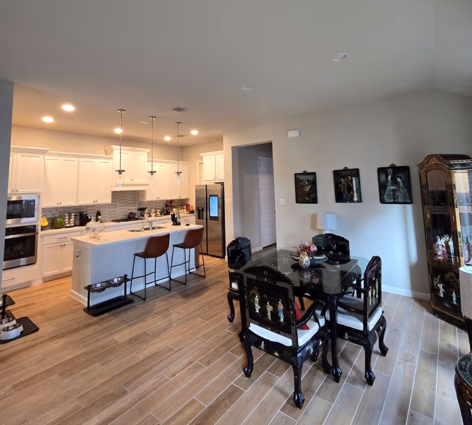A modern kitchen and dining area with white cabinetry, stainless steel appliances, and a kitchen island with two brown bar stools. The dining area features a black dining table with four matching chairs decorated with intricate designs. The floor is covered with wood-like tiles, and the walls are light-colored with three framed pictures and a lamp on a side table.
