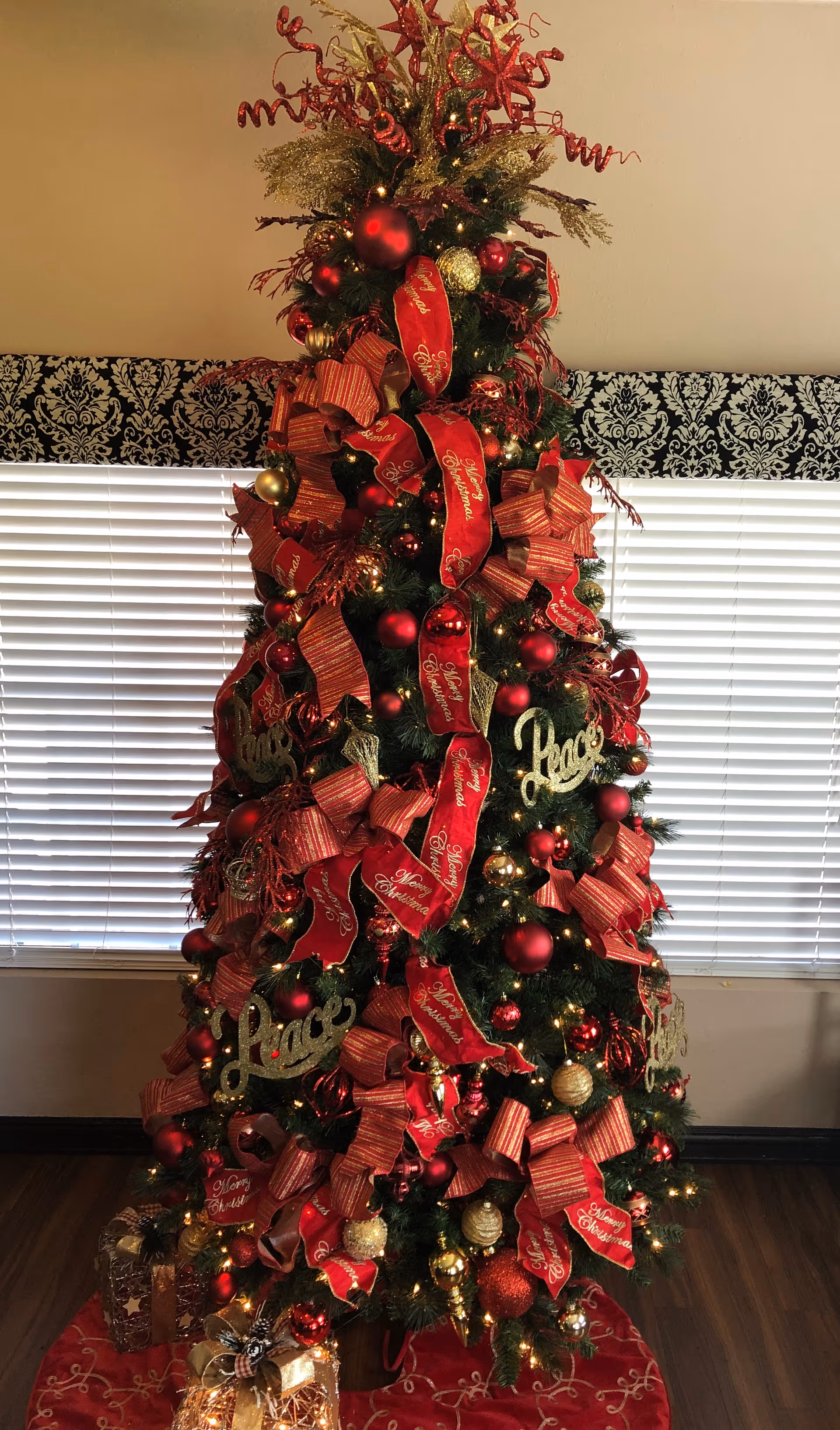 A tall Christmas tree decorated with red and gold ribbons, ornaments, and lights standing in front of a window with blinds.