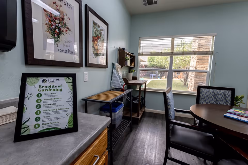 A small room with light blue walls featuring two framed floral prints and a framed sign titled 'Benefits of Gardening' on a countertop. The room has a window with blinds showing an outdoor garden area. There is a round table with patterned chairs, a small wooden shelf with gardening supplies, and a rolling cart with storage bins underneath.