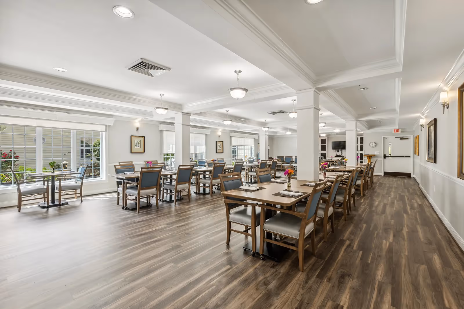 A spacious, well-lit dining room with multiple wooden tables and chairs arranged neatly. The room features large windows allowing natural light to fill the space, light-colored walls, and ceiling lights. There are framed pictures on the walls and small flower arrangements on the tables.