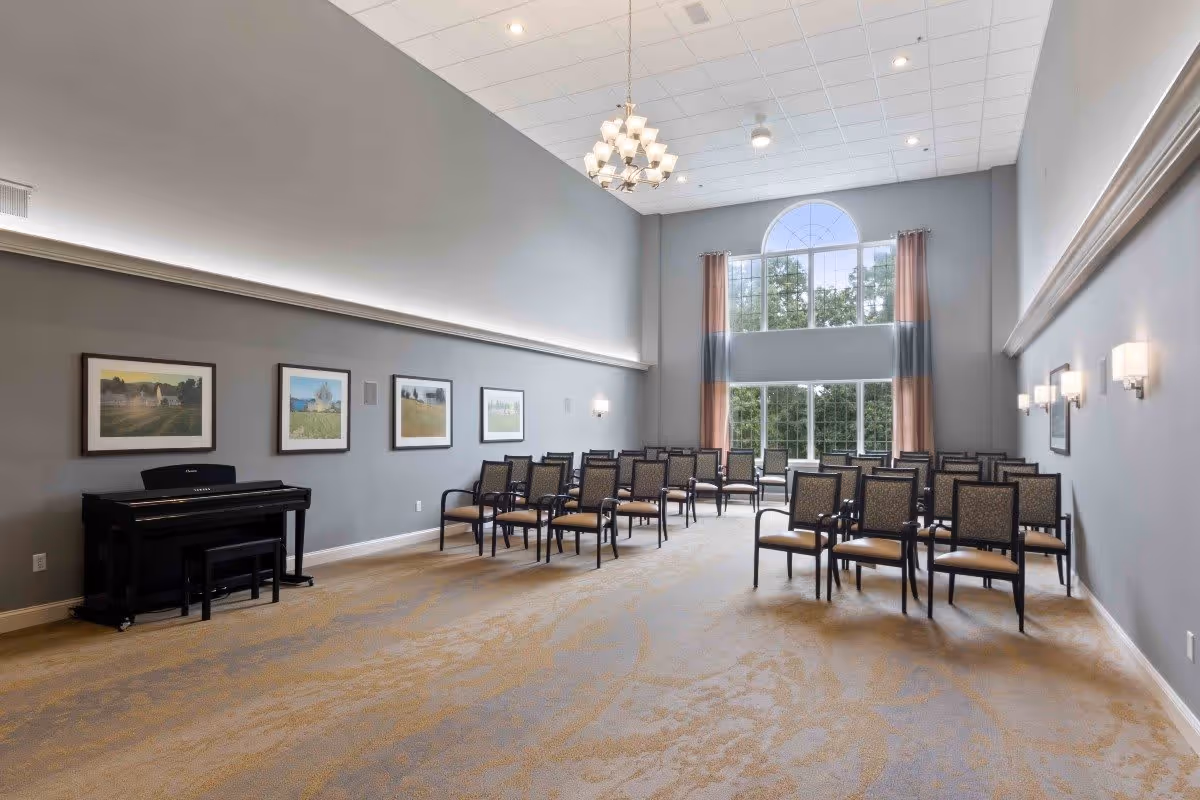 A spacious room with high ceilings and large windows letting in natural light. The room has rows of chairs arranged facing a black piano against a gray wall adorned with framed landscape pictures. There is a chandelier hanging from the ceiling and wall-mounted lights on the right side.