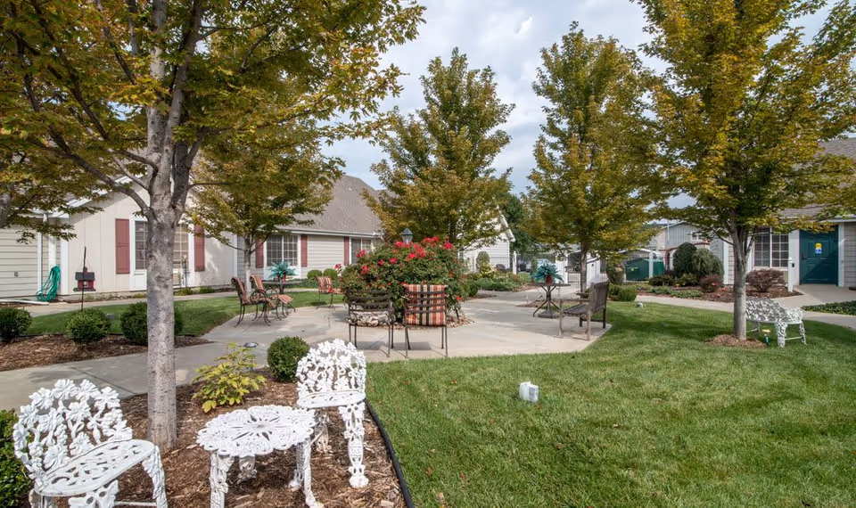 Outdoor courtyard area at a senior living facility with green grass, several trees, and a variety of seating options including white decorative metal chairs and tables, and cushioned chairs around a central flower bed with red flowers. Single-story buildings with beige siding and maroon shutters surround the courtyard under a partly cloudy sky.