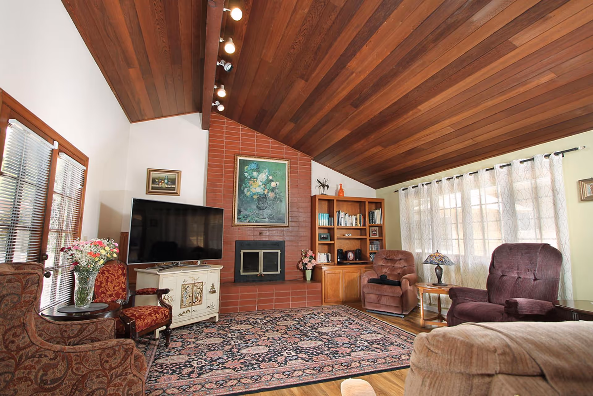 A cozy living room with a wooden vaulted ceiling and a large patterned rug on the floor. The room features a brick fireplace with a floral painting above it, a flat-screen TV on a white cabinet, and a wooden bookshelf filled with books and decorative items. There are several upholstered chairs and a small side table with a lamp near a window with sheer curtains.