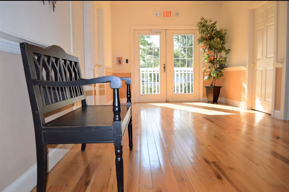 A bright hallway with polished wooden floors, a black wooden bench on the left side, a potted plant on the right near double glass doors with an exit sign above them, and sunlight streaming through the doors.