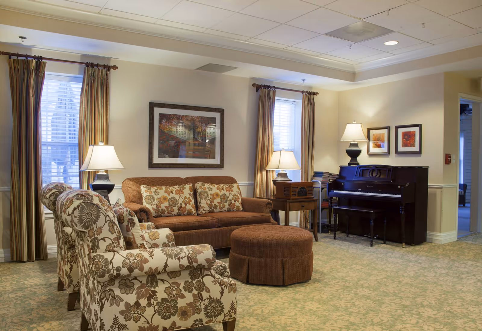 A cozy living room area with a brown floral-patterned sofa and matching armchairs, a round brown ottoman, two table lamps on side tables, a dark upright piano against the wall, framed artwork, and windows with striped curtains.