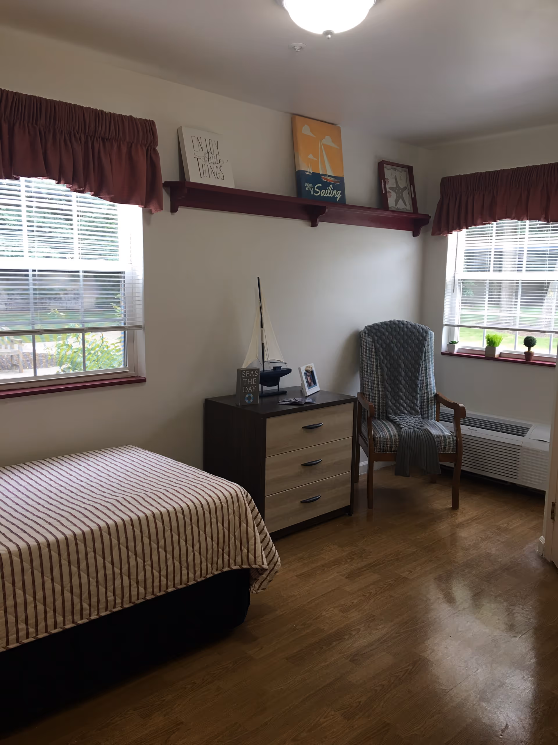 A bedroom with two windows covered by red valances, a bed with a striped bedspread, a wooden dresser with three drawers, a decorative sailboat and framed photos on top, a cushioned armchair with a gray throw draped over it, and a wooden floor.