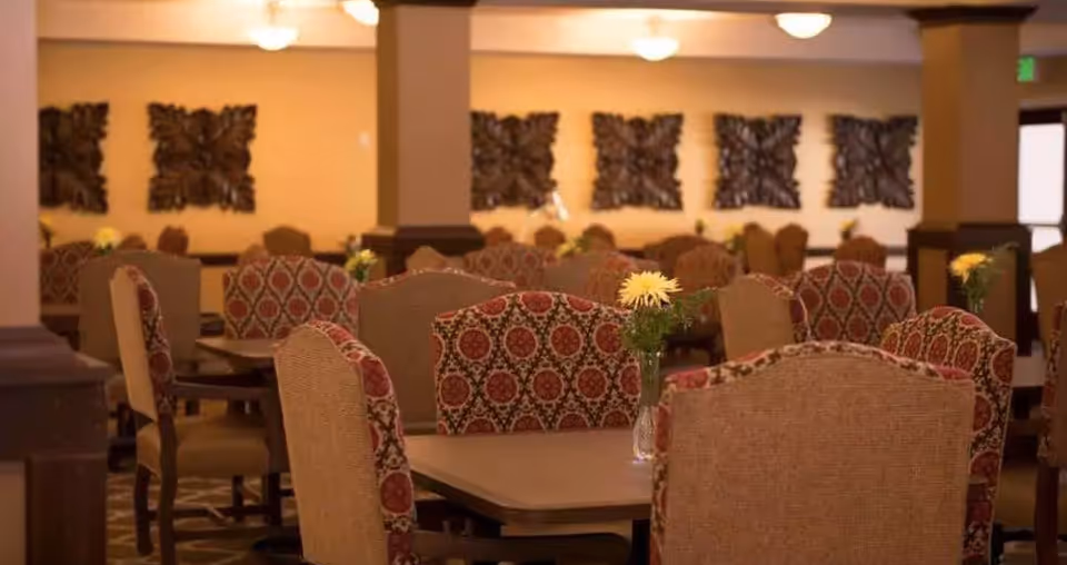 A warm and inviting dining room with multiple tables and cushioned chairs featuring a red and beige patterned upholstery. Each table has a small vase with yellow flowers. The walls are decorated with dark wooden ornamental panels, and the room is softly lit with ceiling lights.