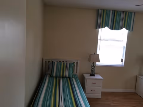 A small bedroom with a single bed against the left wall. The bed has a striped bedspread and matching pillow in shades of green, blue, and white. Next to the bed is a white nightstand with two drawers and a decorative lamp on top. A window with a striped valance matching the bedspread is on the wall above the nightstand. The room has light-colored walls and wooden flooring.