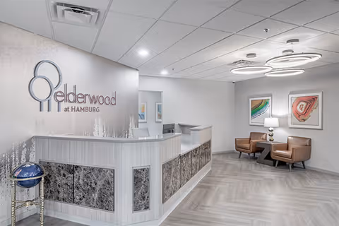 Reception area of Elderwood at Hamburg featuring a modern front desk with marble panels, a globe on a stand to the left, two brown leather chairs with a small table and lamp between them, colorful abstract artwork on the wall, and circular ceiling lights.