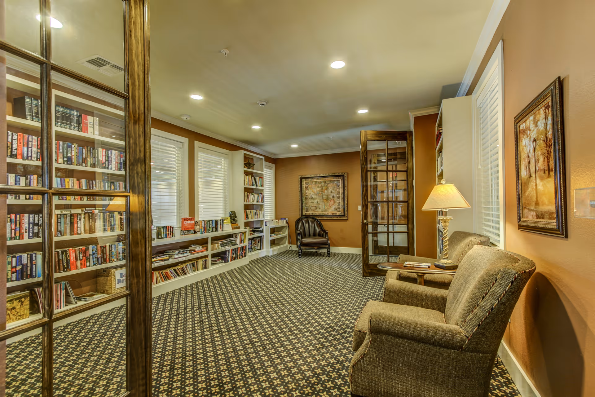 Cozy library-style common room with bookshelves, armchairs, a side table with lamp, and patterned carpet.