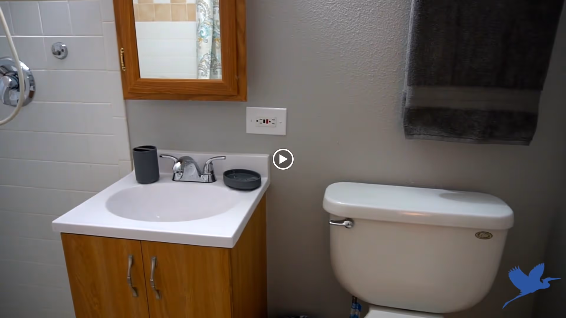 A bathroom with a white sink and wooden cabinet below, a wall-mounted mirror with a wooden frame above the sink, a white toilet next to the sink, a dark gray towel hanging on the wall, and a shower area partially visible on the left side.
