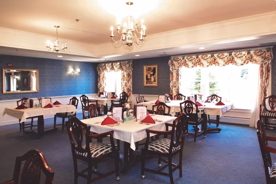 A dining room with multiple tables covered in white tablecloths and set with red folded napkins, menus, and silverware. The room has blue carpet, blue patterned wallpaper, floral curtains on large windows, and chandeliers hanging from the ceiling. Wooden chairs with striped cushions surround the tables.