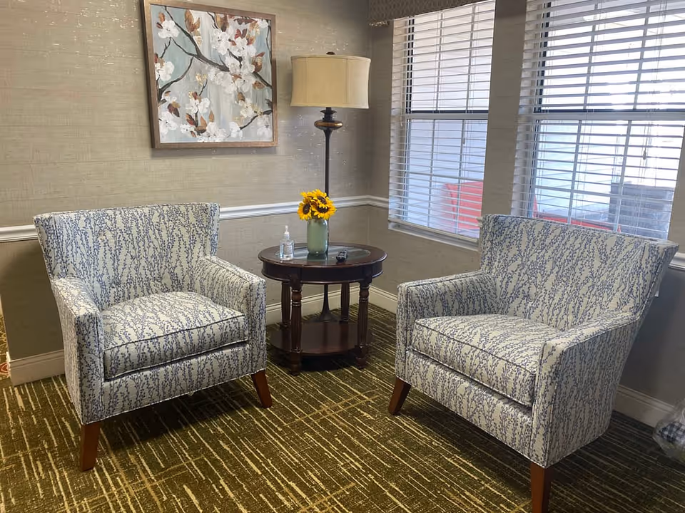 Two patterned armchairs facing each other with a round wooden side table holding a vase of sunflowers and a lamp beside a window.