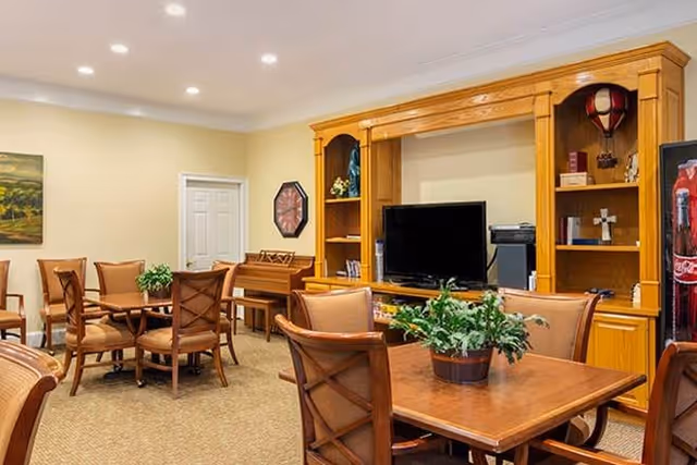 A cozy common area with wooden tables and chairs arranged for seating. There is a large wooden entertainment center with a flat-screen TV and shelves holding decorative items. A piano is visible against the wall, and a potted plant is placed on one of the tables. The room has beige walls and carpeted flooring.