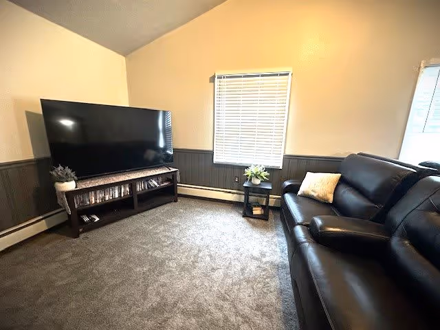 A cozy living room with a large flat-screen TV on a wooden stand, a small side table with a plant and decorative items, and a black leather sofa with a light-colored pillow. The room has carpeted flooring, beige walls with dark wainscoting, and two windows with blinds.