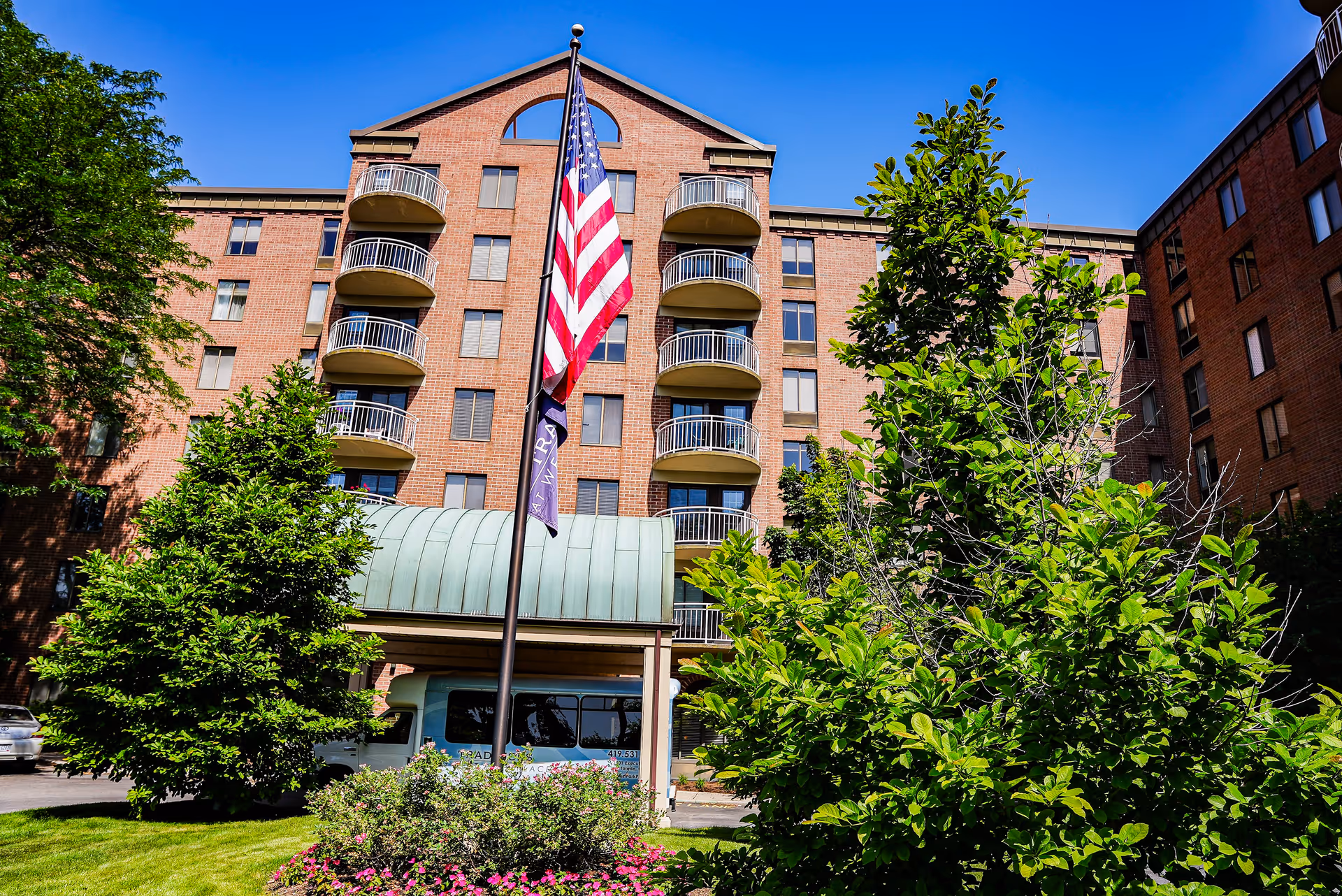 Exterior view of a multi-story brick senior living facility with balconies, an American flag on a flagpole, green trees, and a blue sky.