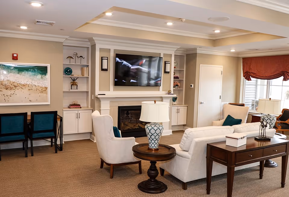 A cozy living room with beige walls and carpet, featuring a white sofa and armchair with teal cushions, a wooden side table with a patterned lamp, a fireplace with a mounted flat-screen TV above it, built-in shelves with decorative items, a desk with two teal chairs, and a large window with red curtains letting in natural light.