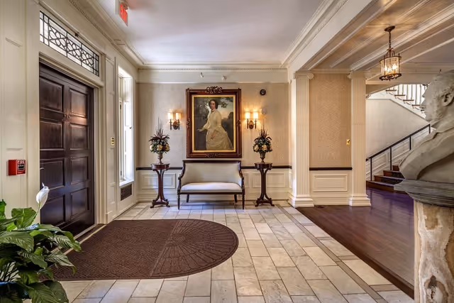 An elegant interior lobby area with a dark wooden door on the left, a beige tiled floor with a brown mat, and a small upholstered bench centered against the wall. Above the bench is a framed portrait of a woman in a white dress, flanked by two wall sconces with lights. Two small round tables with floral arrangements stand on either side of the bench. To the right, there is a bust sculpture on a pedestal and a staircase with a black railing leading upstairs. The walls are decorated with light-colored wallpaper and white wainscoting.