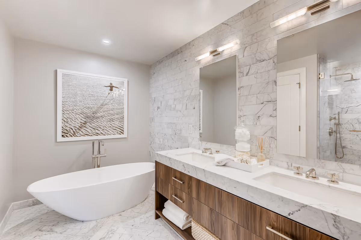 Modern bathroom with a freestanding white bathtub, a double sink vanity with marble countertop and wooden drawers, a large mirror above the sinks, marble tiled walls and floor, and a framed picture of a person on a sandy surface hanging on the wall.