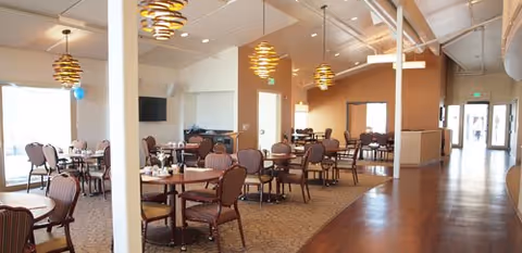 Spacious dining room with multiple round tables and chairs arranged neatly. The room features warm lighting with modern hanging light fixtures, carpeted and hardwood flooring, and large windows allowing natural light. There is a TV mounted on the wall and a doorway leading to another area.