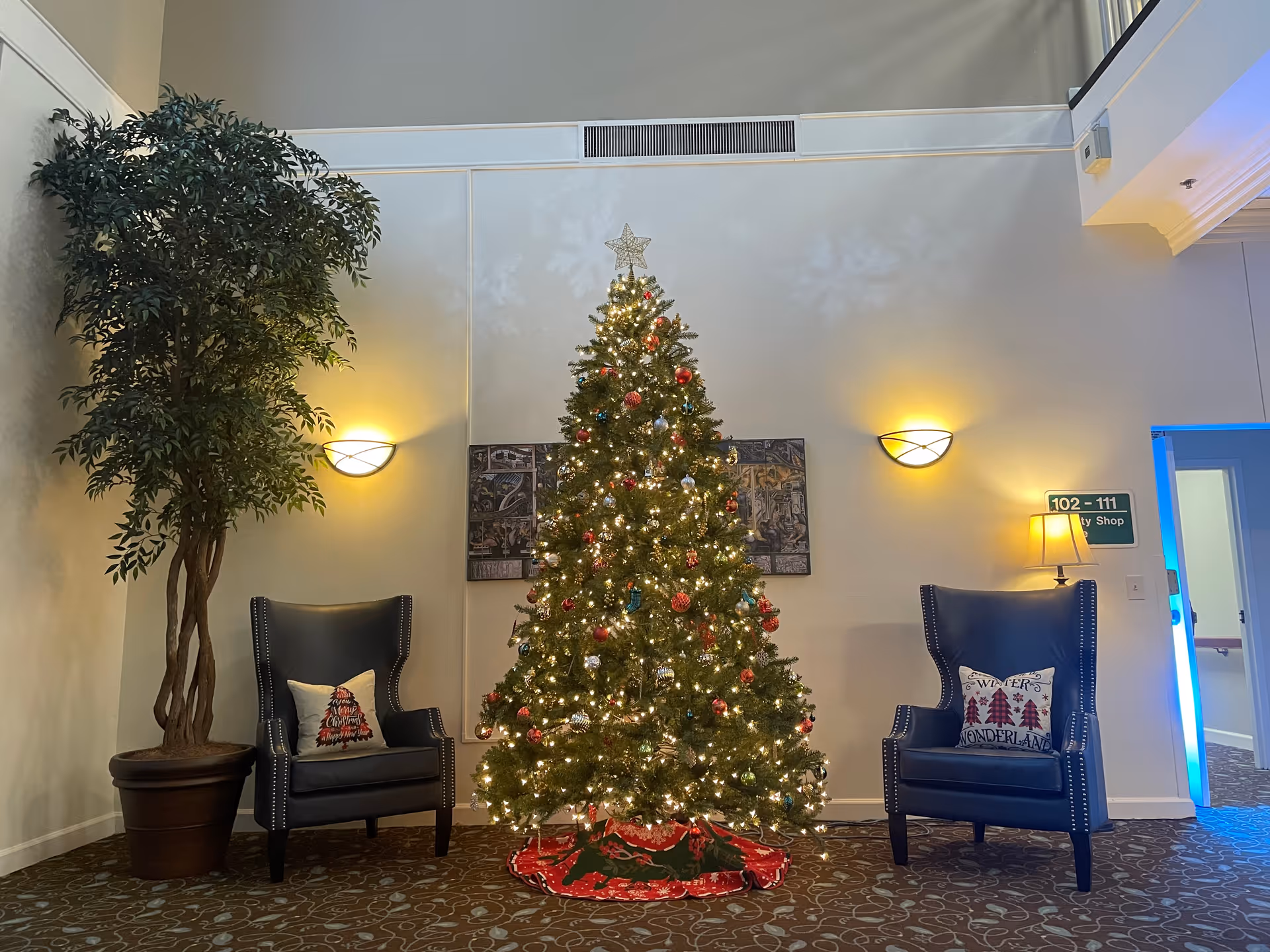 A decorated Christmas tree with lights and ornaments stands in the center of a room. On either side of the tree are two dark blue armchairs with festive pillows. A large potted plant is on the left side, and a small lamp on a table is on the right. The walls are light-colored with two wall sconces providing warm lighting. A sign on the right wall indicates room numbers and a shop.