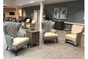A spacious and well-lit common area with comfortable seating including two patterned armchairs, a beige armchair, and a small wooden side table in the center. The room has a neutral color palette with gray walls, carpeted flooring, and decorative artwork on the wall. In the background, there is a dining area with tables and chairs.