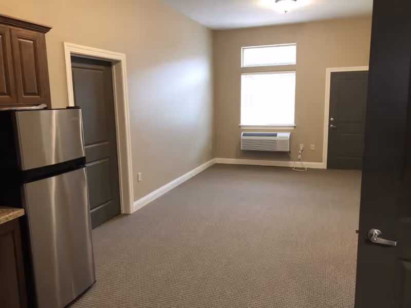 Empty room with beige walls and carpeted floor, a stainless steel refrigerator on the left, two closed gray doors, a window with a white air conditioning unit below it, and a ceiling light fixture.