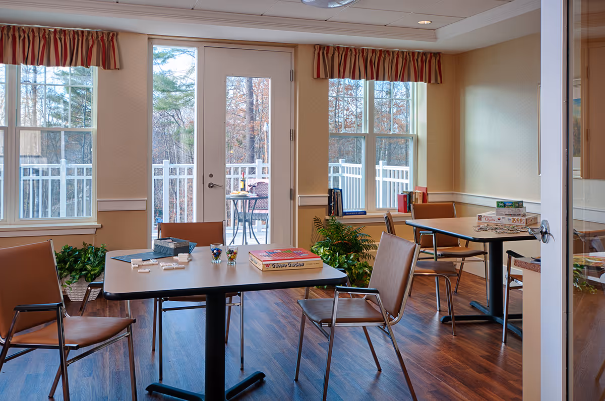 A bright room with large windows and a glass door leading to an outdoor patio. Inside, there are two tables with brown chairs around them. On the tables are board games and puzzle boxes. The room has wood flooring, beige walls, and plants near the windows. Outside, there is a small table with wine and glasses on the patio.