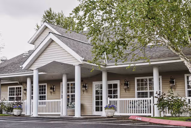 Exterior view of a senior living facility building with beige siding, white columns, and a covered porch area. There are several windows and a door visible, along with potted plants and a tree partially covering the right side of the image.