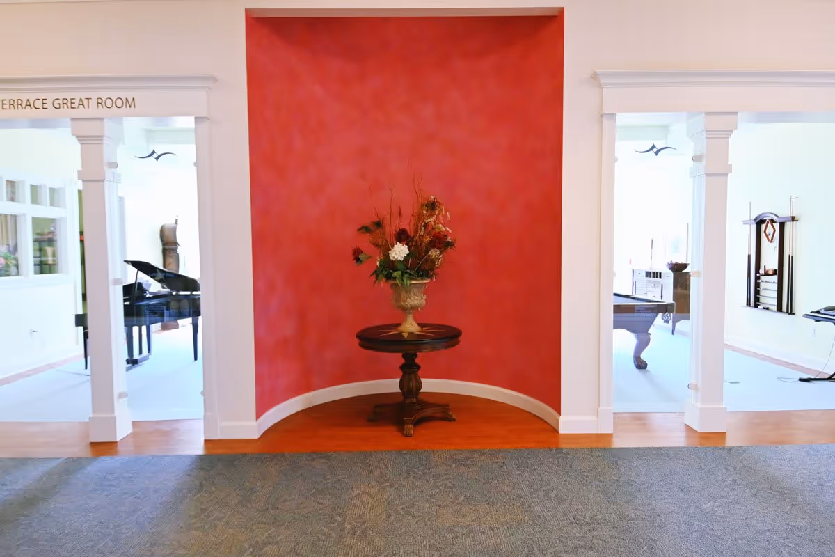 Interior view of a senior living facility with a red accent wall featuring a round wooden table holding a floral arrangement. On either side of the wall are open doorways leading to a bright room with a grand piano on the left and a pool table on the right. The floor has a combination of carpet and wood.
