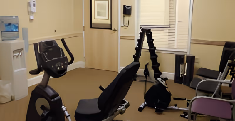 A small exercise room with two stationary exercise bikes, several chairs along the wall, a water cooler, and a door with a window. The walls are painted beige and there is a window with blinds.