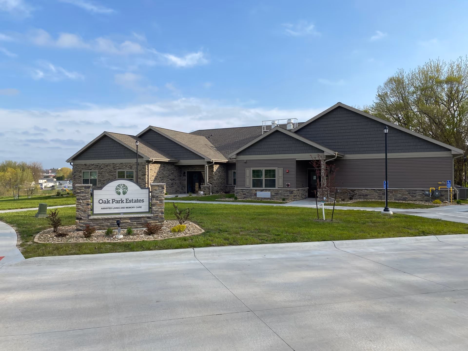Exterior front view of Oak Park Estates Assisted Living and Memory Care building with a stone and siding facade, a manicured lawn, and a sign displaying the facility name in front.