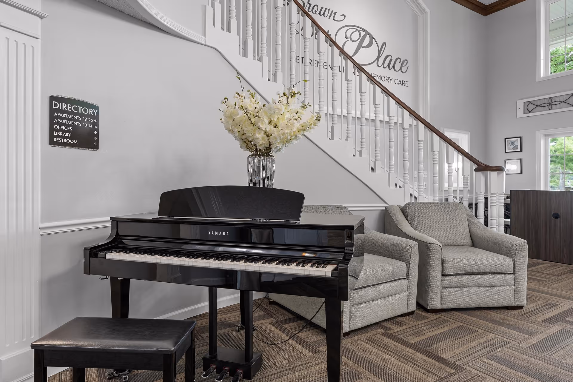 Interior view of a senior living facility lobby area featuring a black Yamaha piano with a vase of white flowers on top, two gray upholstered armchairs, a staircase with white railings, and a directory sign on the wall indicating locations of apartments, offices, library, and restroom. The wall behind the staircase has the text 'Brown Deer Place Retirement & Memory Care'.