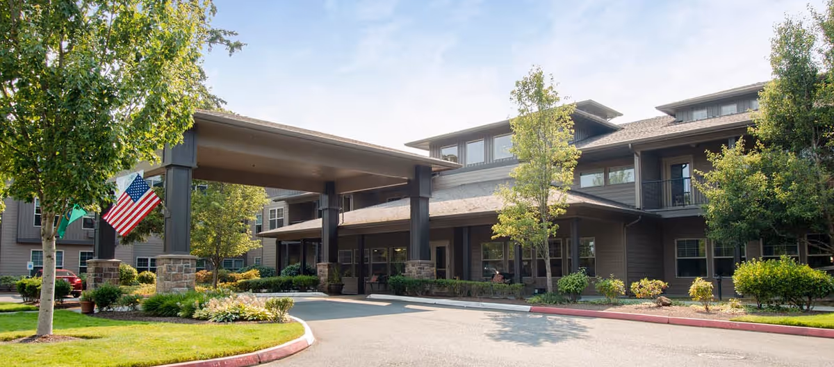 Front exterior of a senior living facility with a covered porte-cochère entrance, landscaped grounds, and an American flag.